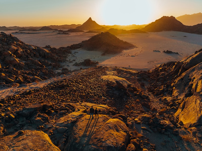 Two people standing on a rocky desert plateau at sunset with distant mountain ranges
