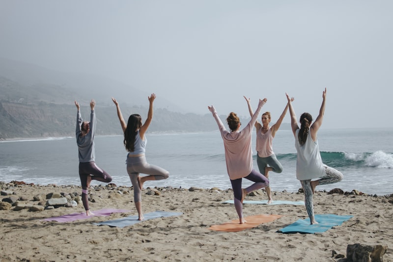 Five women practicing tree pose yoga on mats on a sandy beach facing the ocean